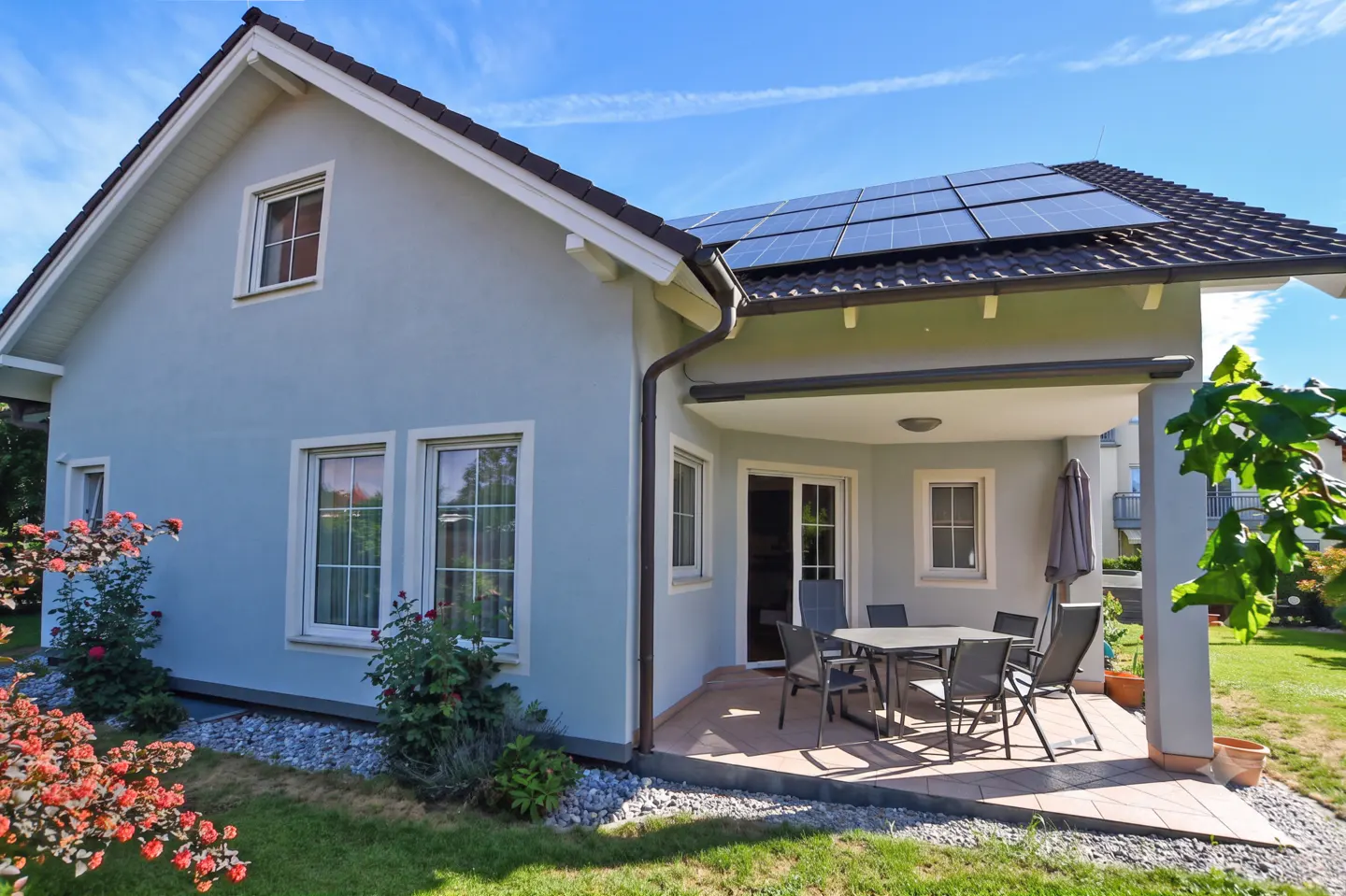 Light blue house with solar panels on the roof, featuring a covered patio with a dining set and a garden in the foreground.
