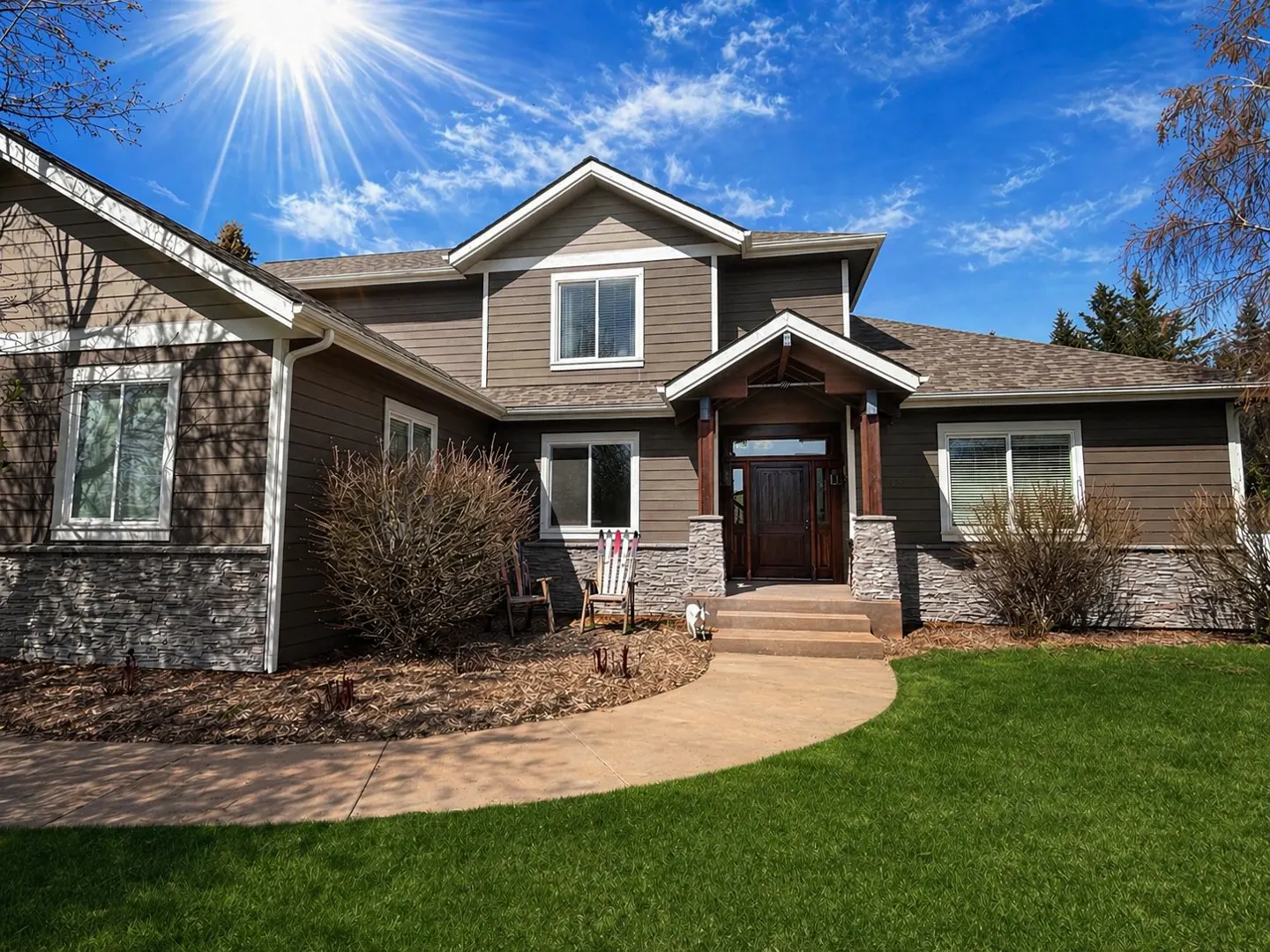 Two-story brown house with stone accents, a curved walkway, and green lawn under a bright blue sky.