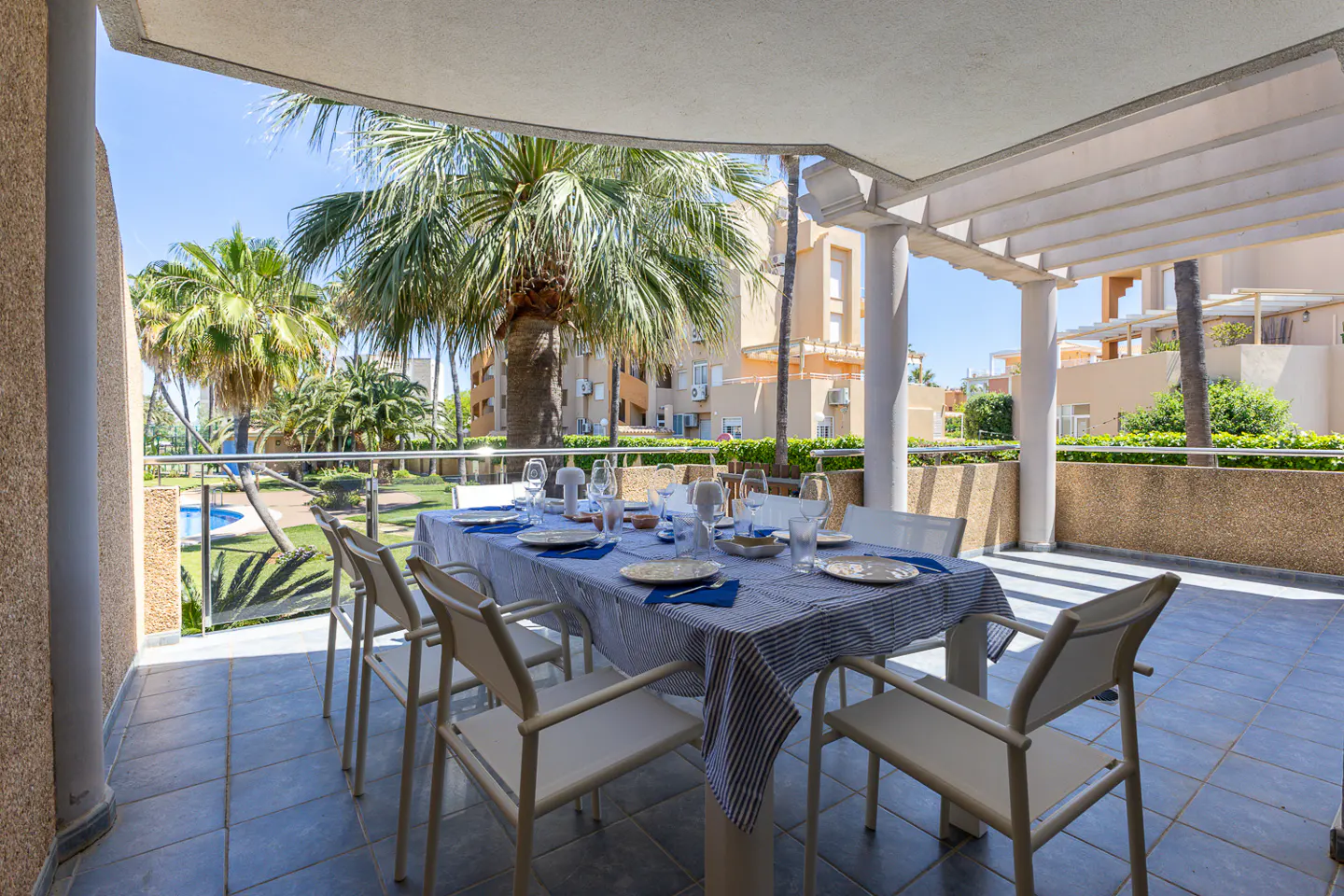 Outdoor dining area with a table set for a meal, featuring a blue and white striped tablecloth, plates, glasses, and chairs. Palm trees and buildings are visible in the background.