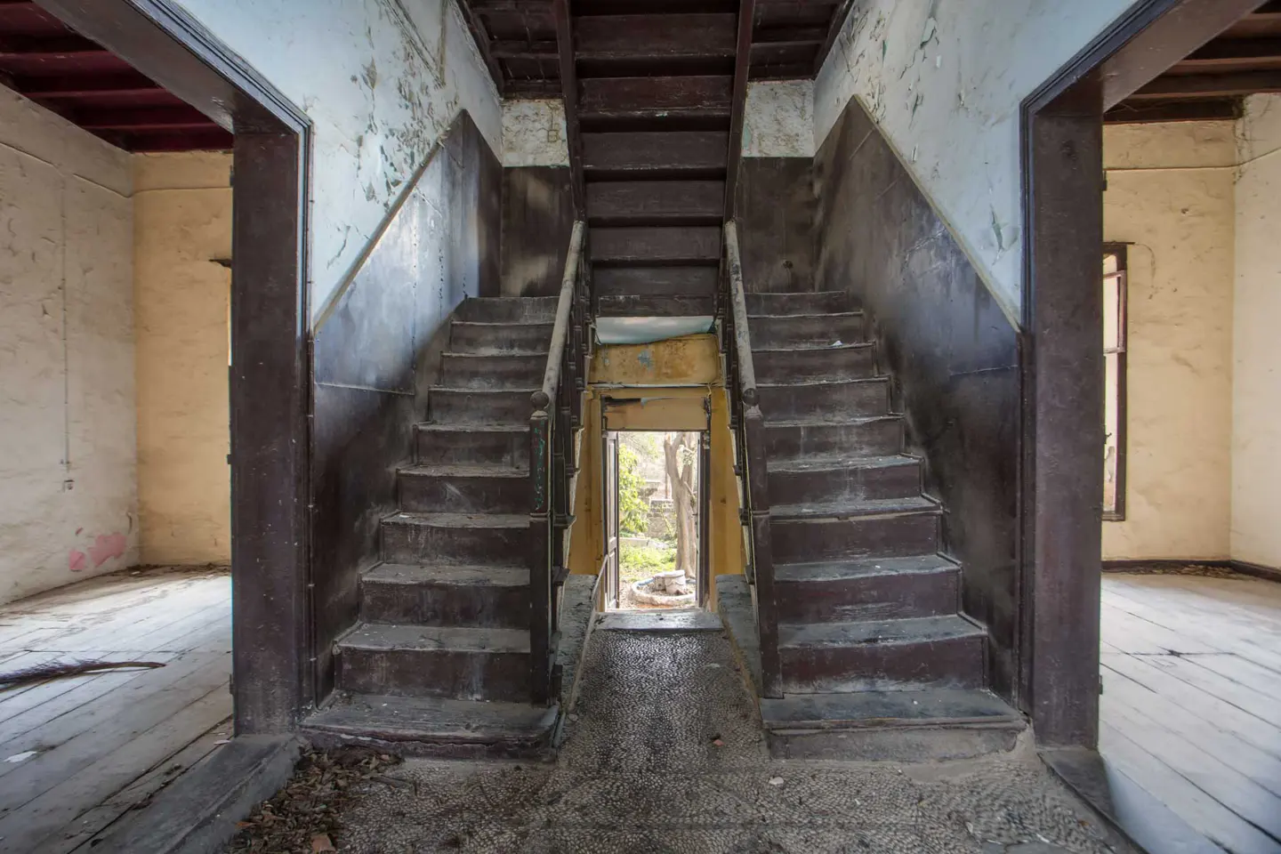 Interior view of a dilapidated building featuring a central staircase with peeling paint and a doorway leading to a garden.