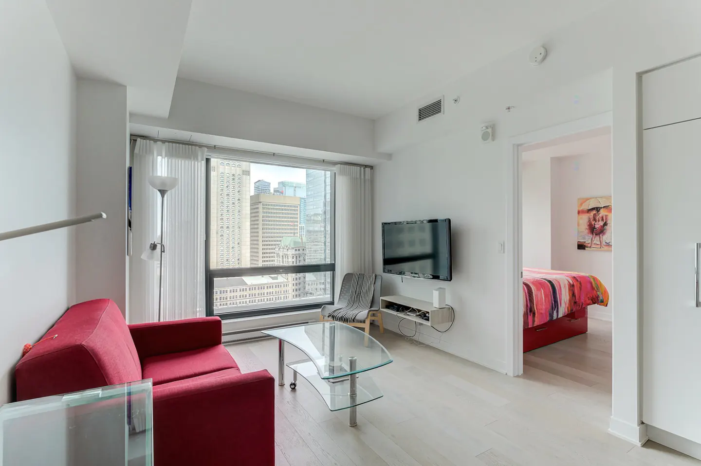 Bright, modern apartment living room with a red sofa, glass table, and city view. A bedroom is visible through an open doorway.