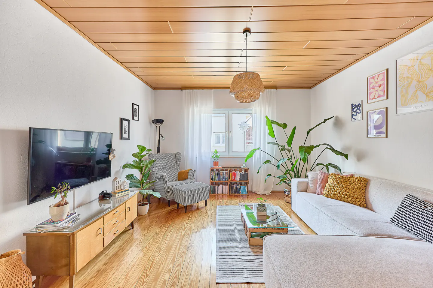 Bright living room with wood floors, white walls, and a wood plank ceiling. A white sofa, gray chair, and plants fill the space.