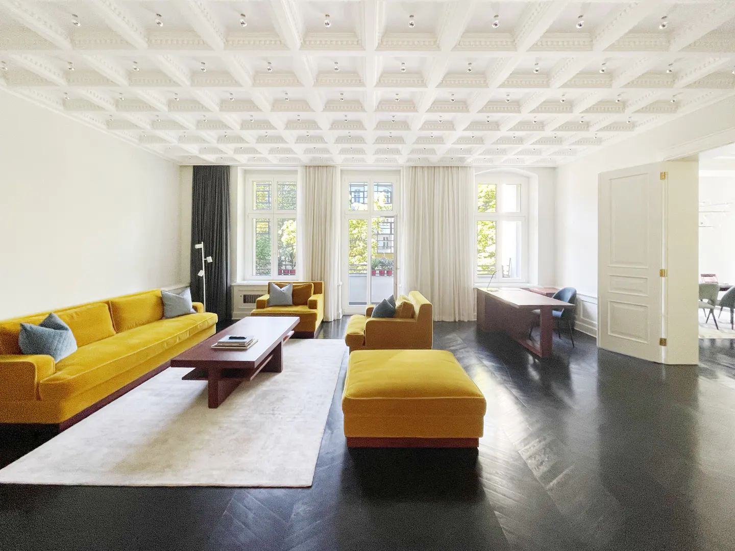 Bright living room with yellow velvet sofa, chairs, and ottoman. White walls, coffered ceiling, dark wood floors, and large windows.