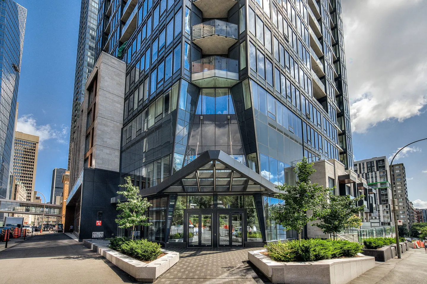 Modern glass high-rise building with a covered entrance and small trees. Other buildings are visible in the background.