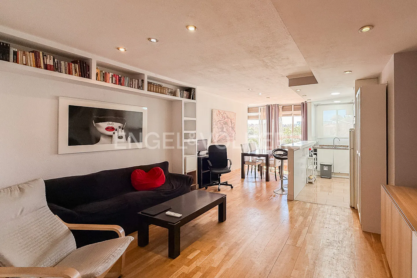 Bright, open-concept living space with hardwood floors. Black sofa with red heart pillow, art, dining table, and kitchen visible.