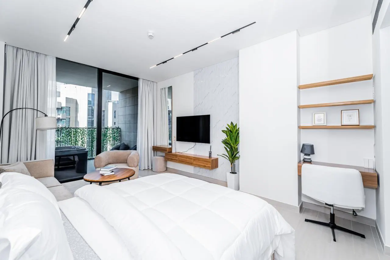 Bright, modern bedroom with a white bed, desk, TV, and balcony with city views. Neutral tones and clean lines create a serene atmosphere.