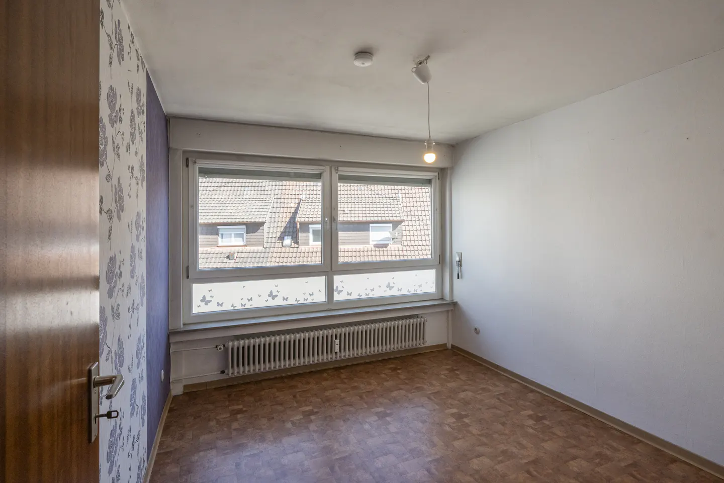 Empty room with wood floor, white walls, and a large window showing rooftops. A radiator sits beneath the window. A brown door is on the left.
