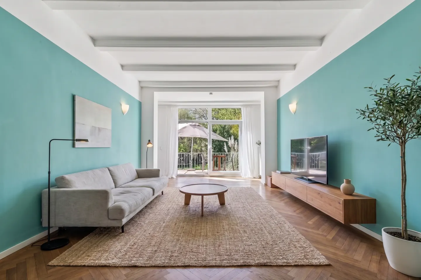 Bright living room with aqua walls, white trim, and wood floors. A beige sofa, round table, and TV stand furnish the space. Balcony visible through glass doors.