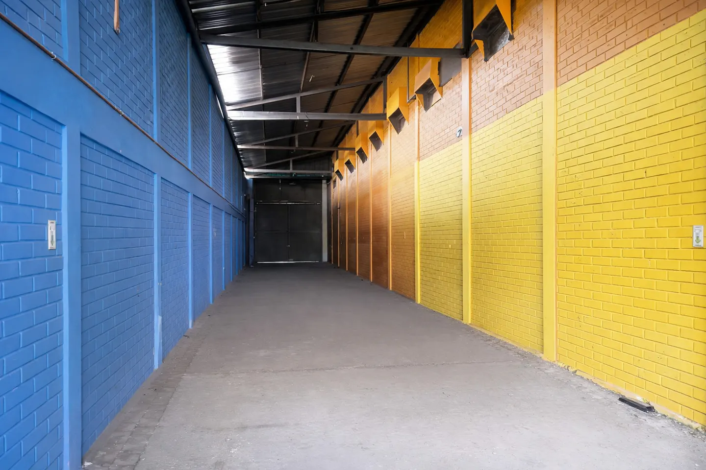 Long, empty warehouse corridor with blue and yellow brick walls and a concrete floor leading to a dark doorway.