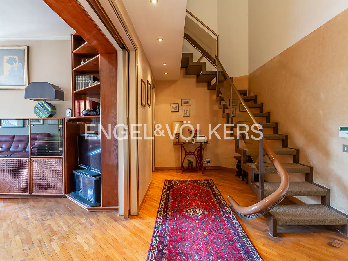 Interior view showing a wooden staircase with a curved railing, a red patterned rug, and a living area with bookshelves.