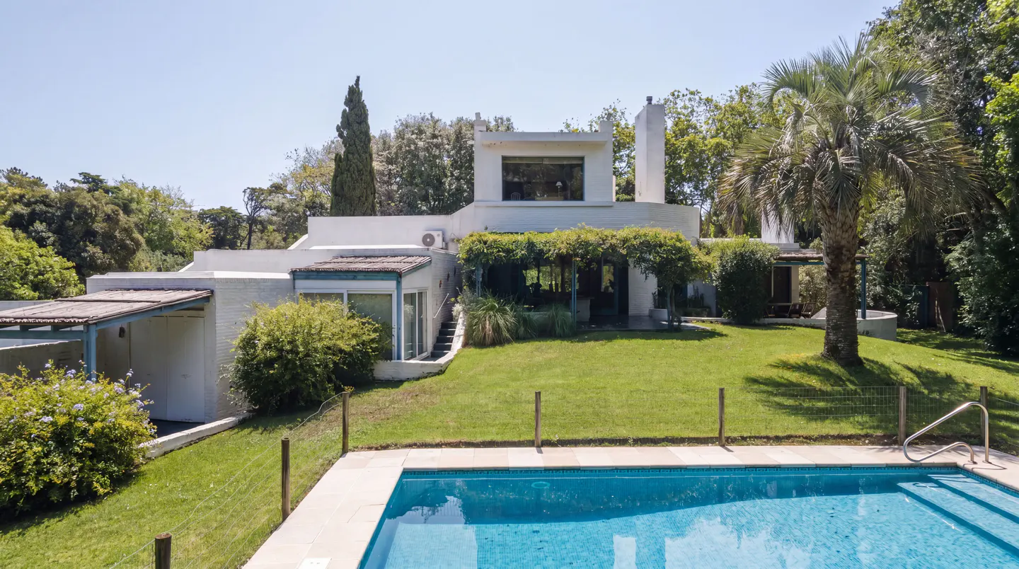 A white house with a blue pool in the foreground and green trees in the background.