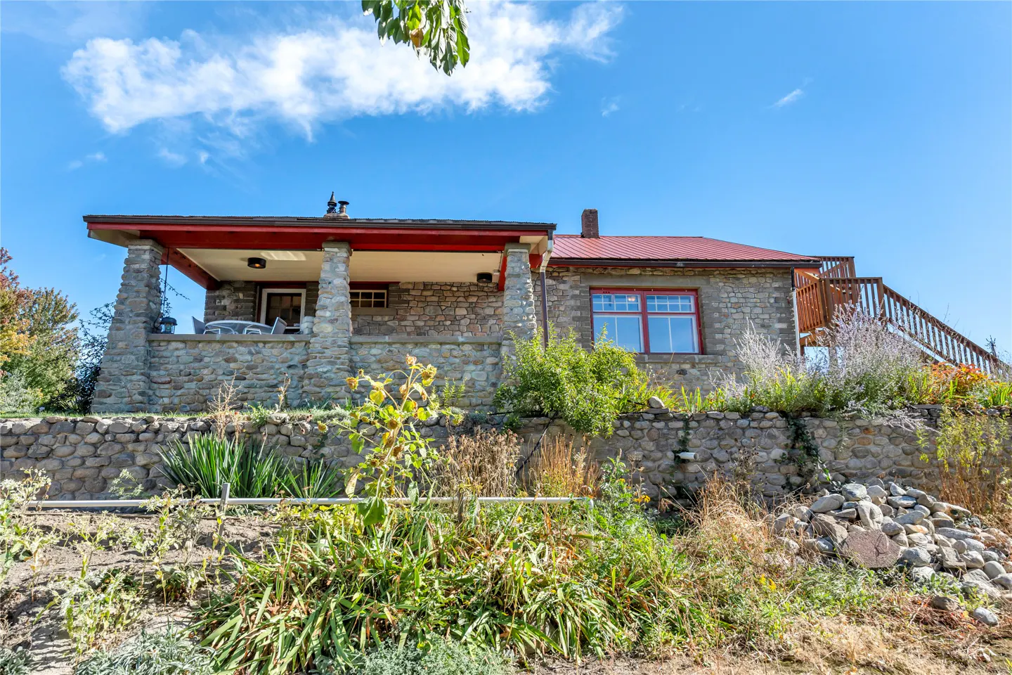 Stone house with red roof and porch, viewed from below on a sunny day. Wooden stairs lead to the roof.