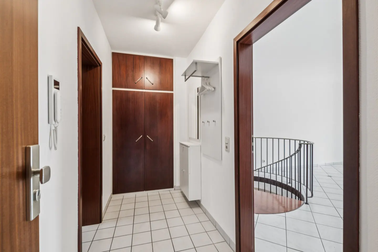 Bright hallway with white tiled floors, dark wood cabinets, a coat rack, and a view of a spiral staircase.