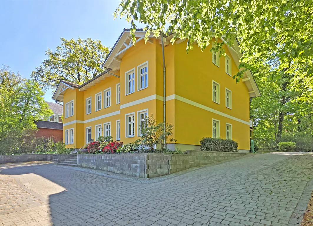 Bright yellow two-story house with white trim, set on a paved driveway surrounded by lush green trees and shrubs.