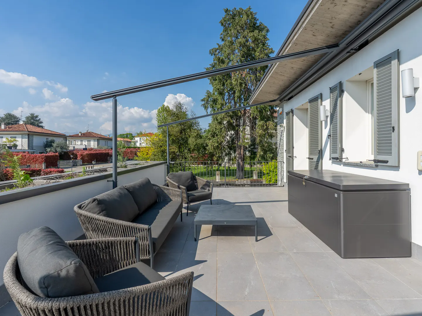 Outdoor patio with gray wicker furniture, a coffee table, and a storage box on a gray tiled floor. White building with gray shutters and a blue sky in the background.