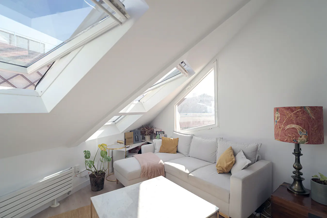 Bright attic living room with white walls, skylights, and a large window. A white sectional sofa with yellow pillows sits near a patterned lamp and wooden chest.