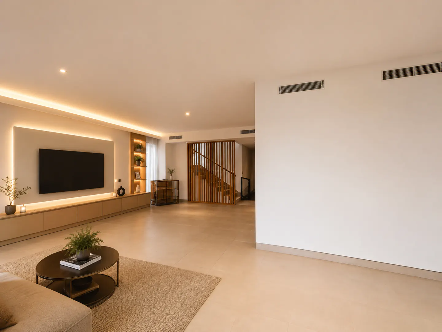 Living room with a TV, a round coffee table with a plant, and a staircase with wooden slats. The walls are white and the floor is beige.