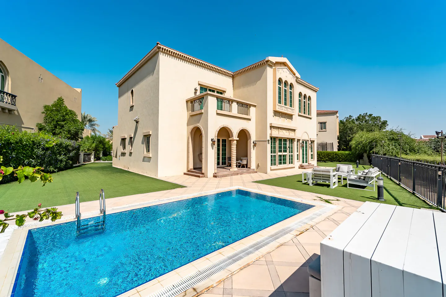 Backyard view of a two-story beige house with a blue swimming pool, green lawn, and white outdoor furniture.