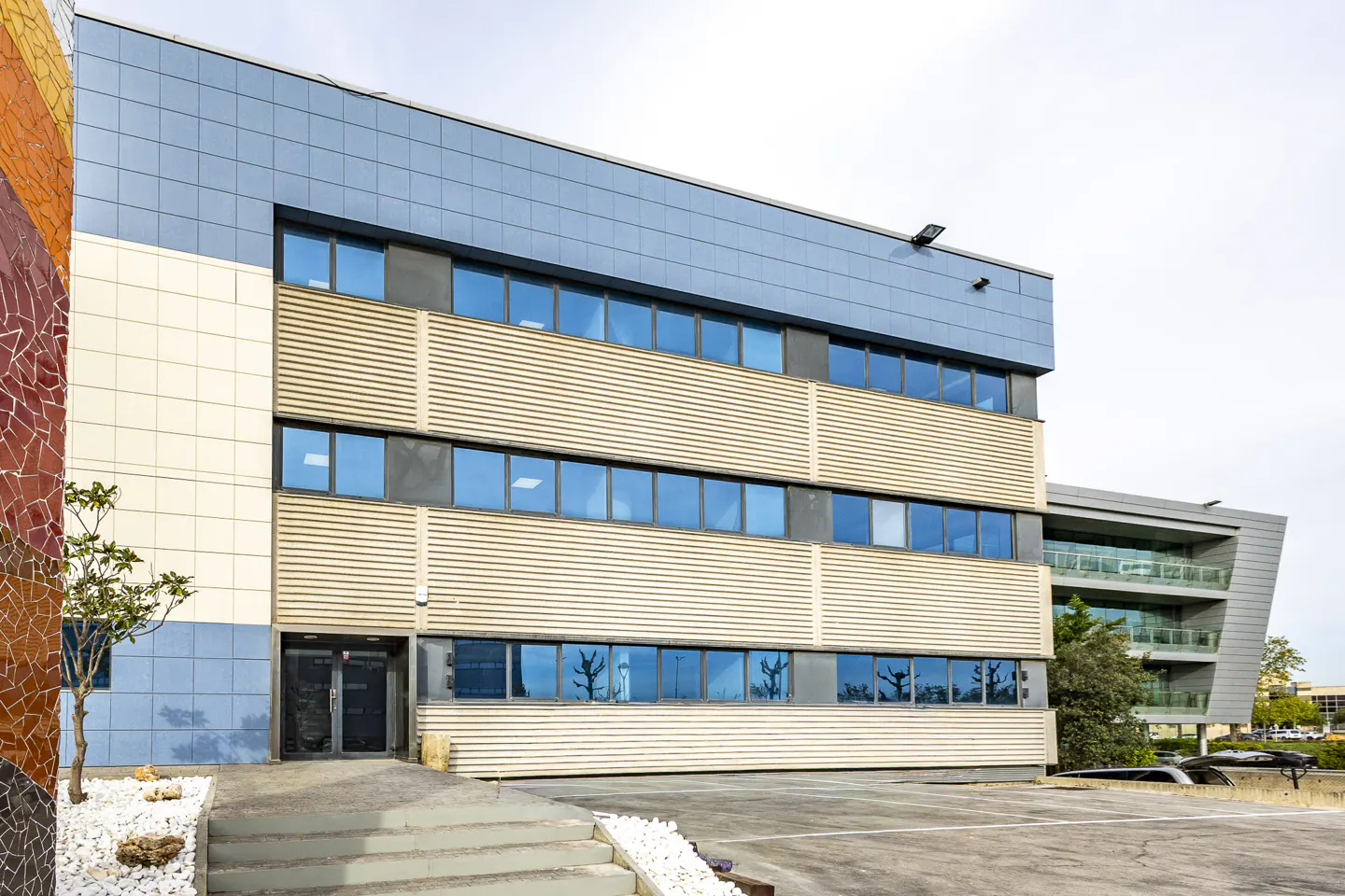 Exterior of a modern, three-story office building with blue windows and a light blue top floor. A mosaic wall is on the left.