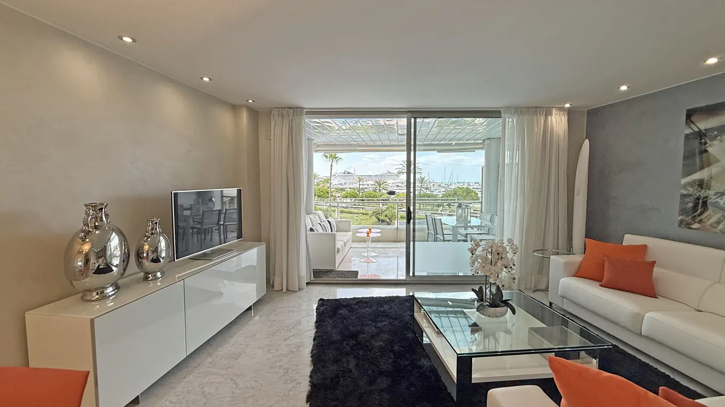 Bright living room with white sofas, orange pillows, glass table, and a balcony overlooking a marina.