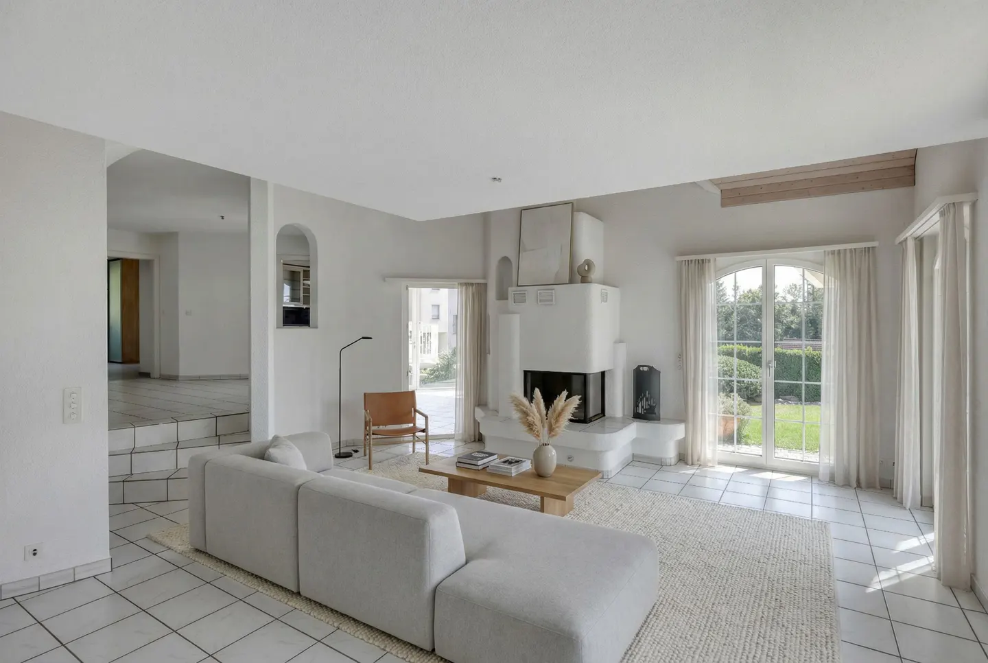 Bright living room with white tile floors, a white couch, and a white fireplace. Large windows with white curtains look out onto a green lawn.