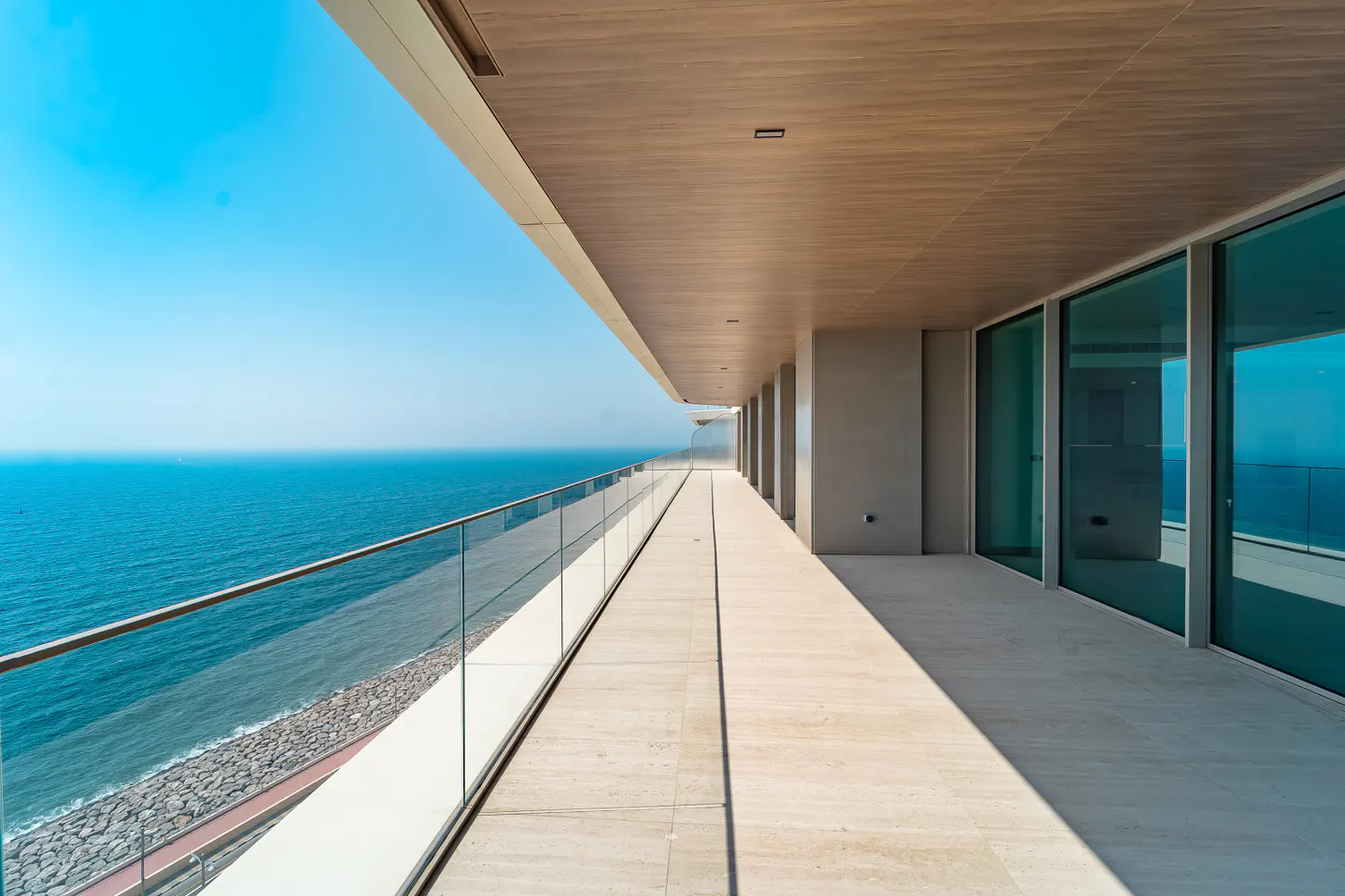 A long, covered balcony with a glass railing overlooks the ocean on a sunny day.