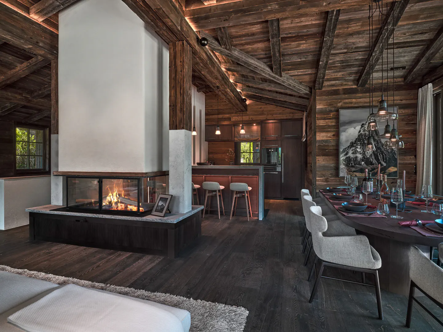 Rustic open-plan living area with a central glass fireplace, wooden beams, dining table, and modern kitchen bar stools.