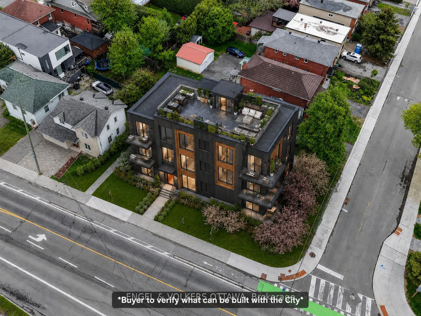 Aerial view of a modern, dark gray apartment building with balconies and a rooftop terrace, surrounded by green trees and residential houses.
