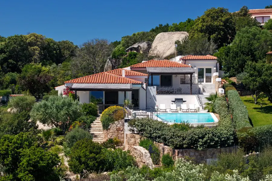 Luxury villa with a red tile roof, a pool, and lush green landscaping on a sunny day.