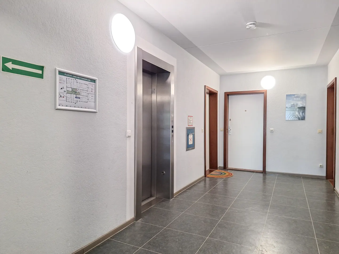Bright apartment building hallway with a metal elevator, white walls, dark tiled floor, and several wooden door frames.