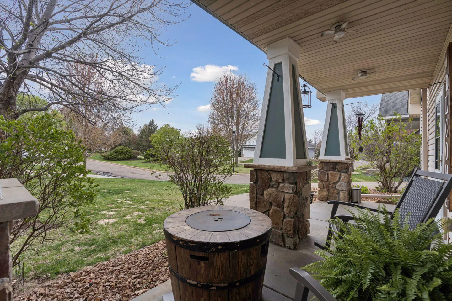 Covered porch with stone pillars, barrel table, and chairs. Green lawn and trees are visible. Blue sky with clouds.