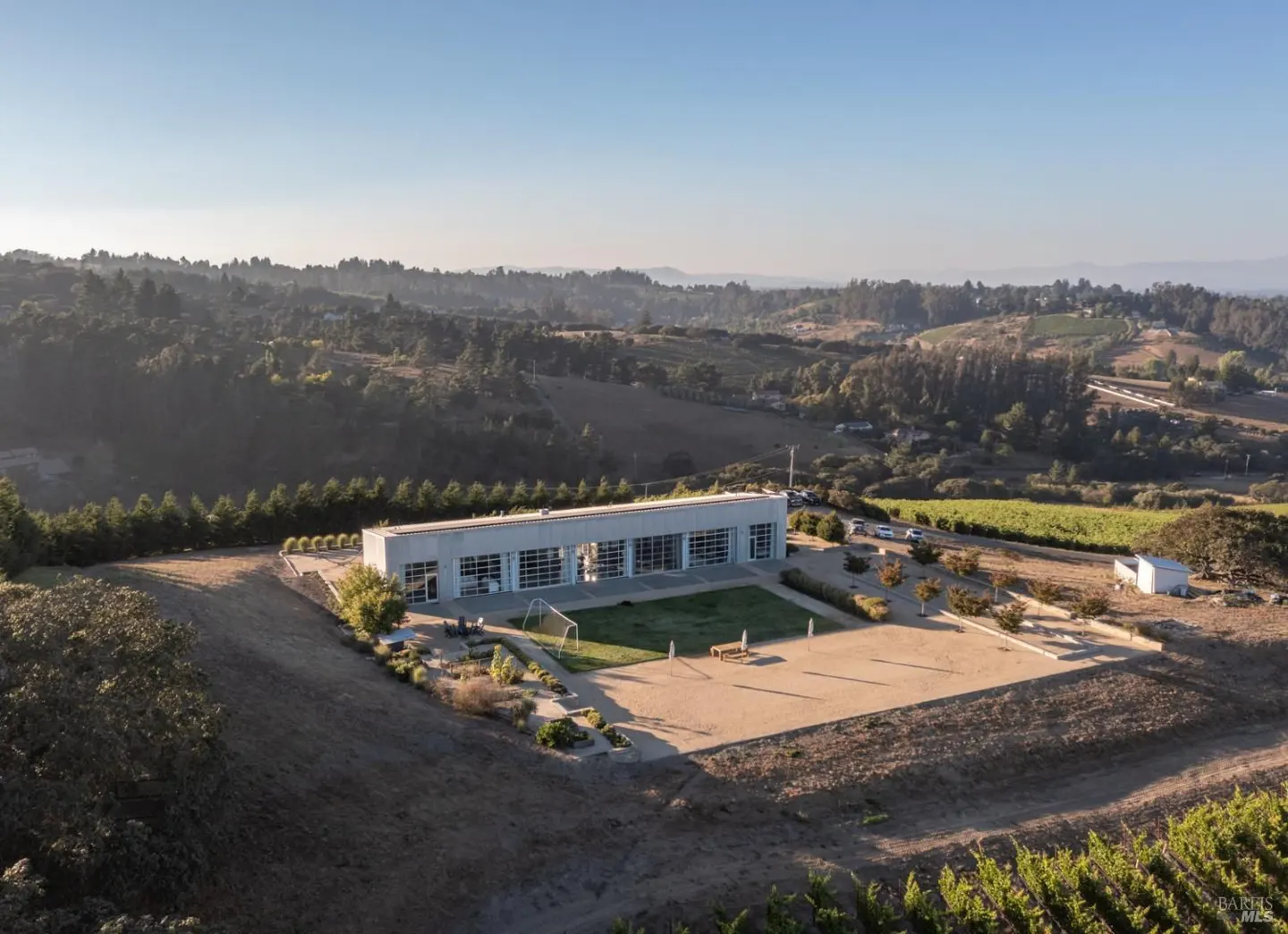 Aerial view of a modern white building with large windows, a green lawn, and a sand court, set against a backdrop of rolling hills and vineyards.