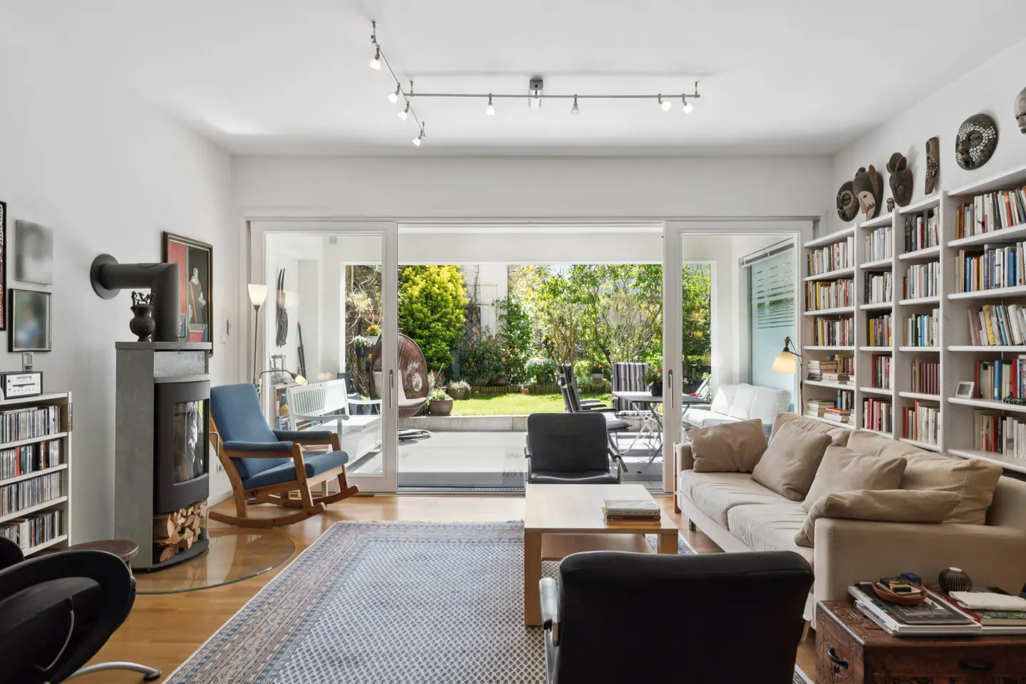 Bright living room with a beige sofa, wood floors, and a large bookshelf. Sliding glass doors open to a green backyard.