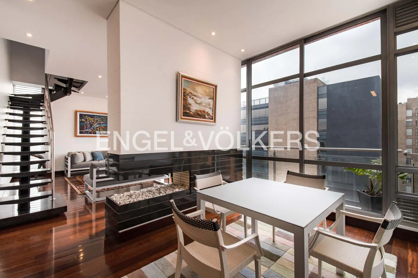Modern living room with a white table, chairs, and a black fireplace. Large windows offer a city view. A staircase leads to an upper level.
