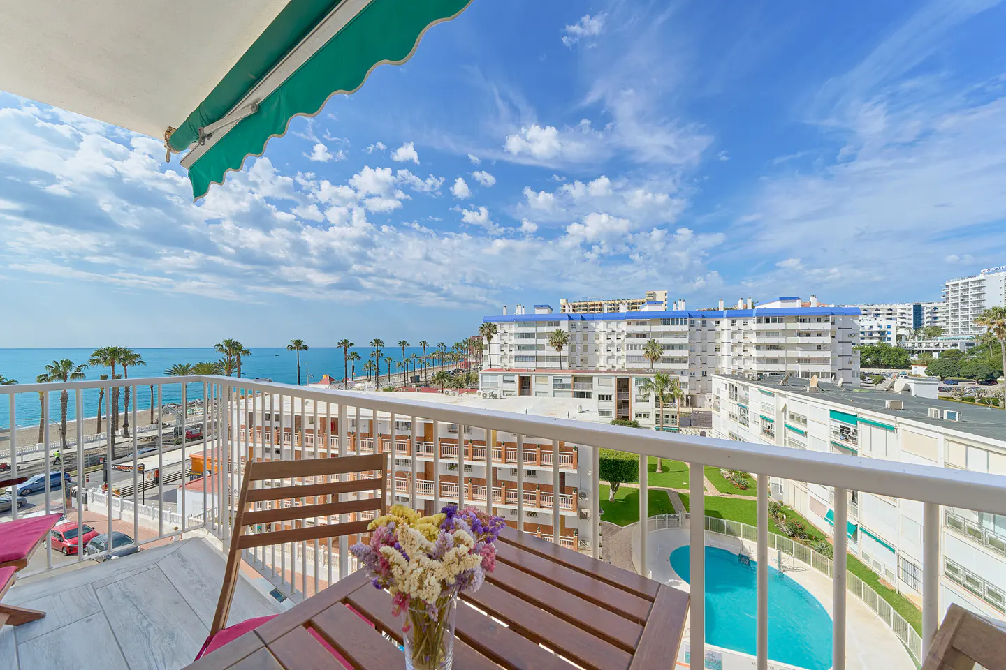 Balcony view with table, chairs, and flowers overlooking the ocean, beach, and city buildings under a blue sky.