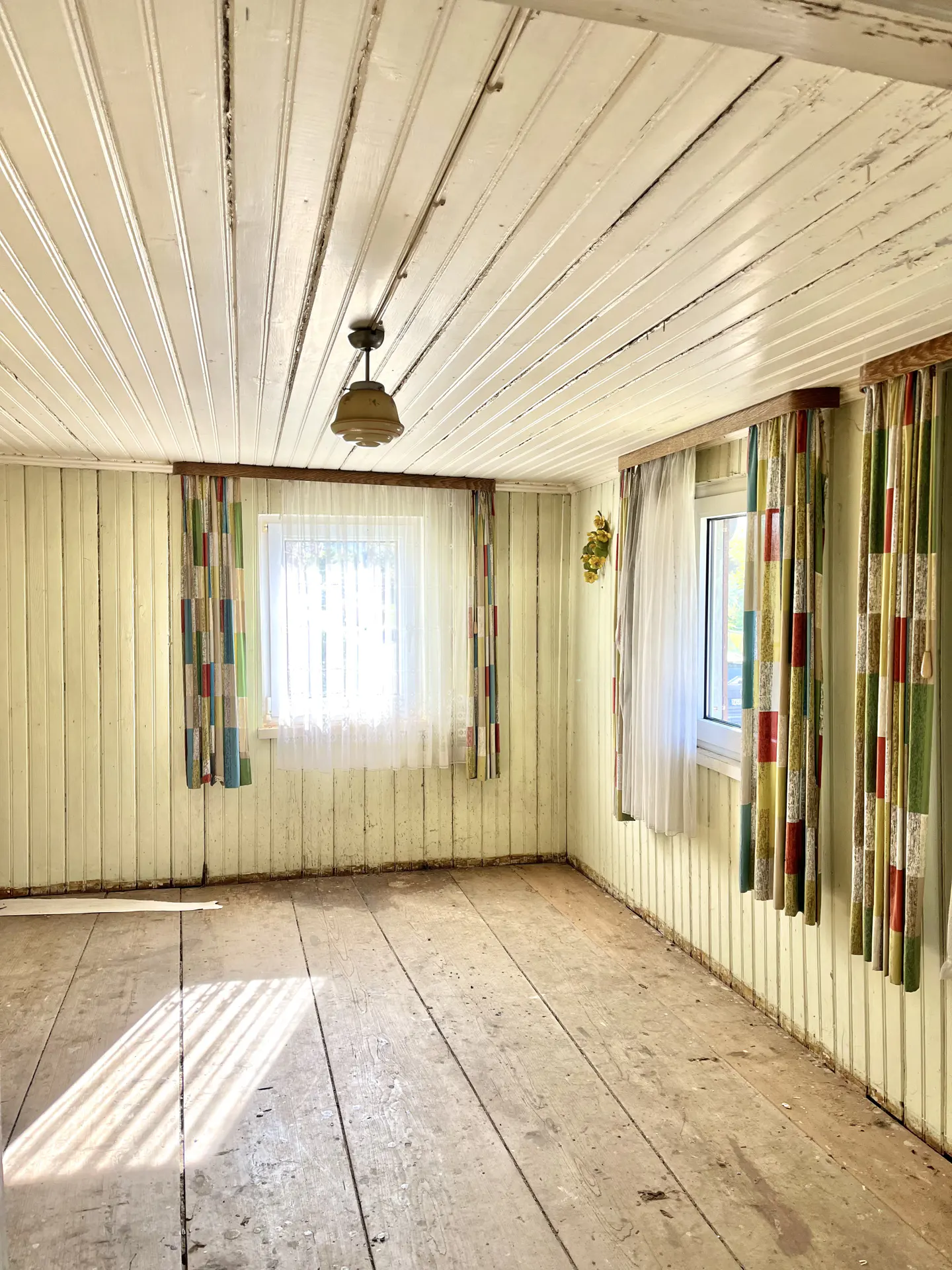 Empty room with wood floors, paneled walls, and a white paneled ceiling. Windows have sheer curtains and colorful patterned drapes.