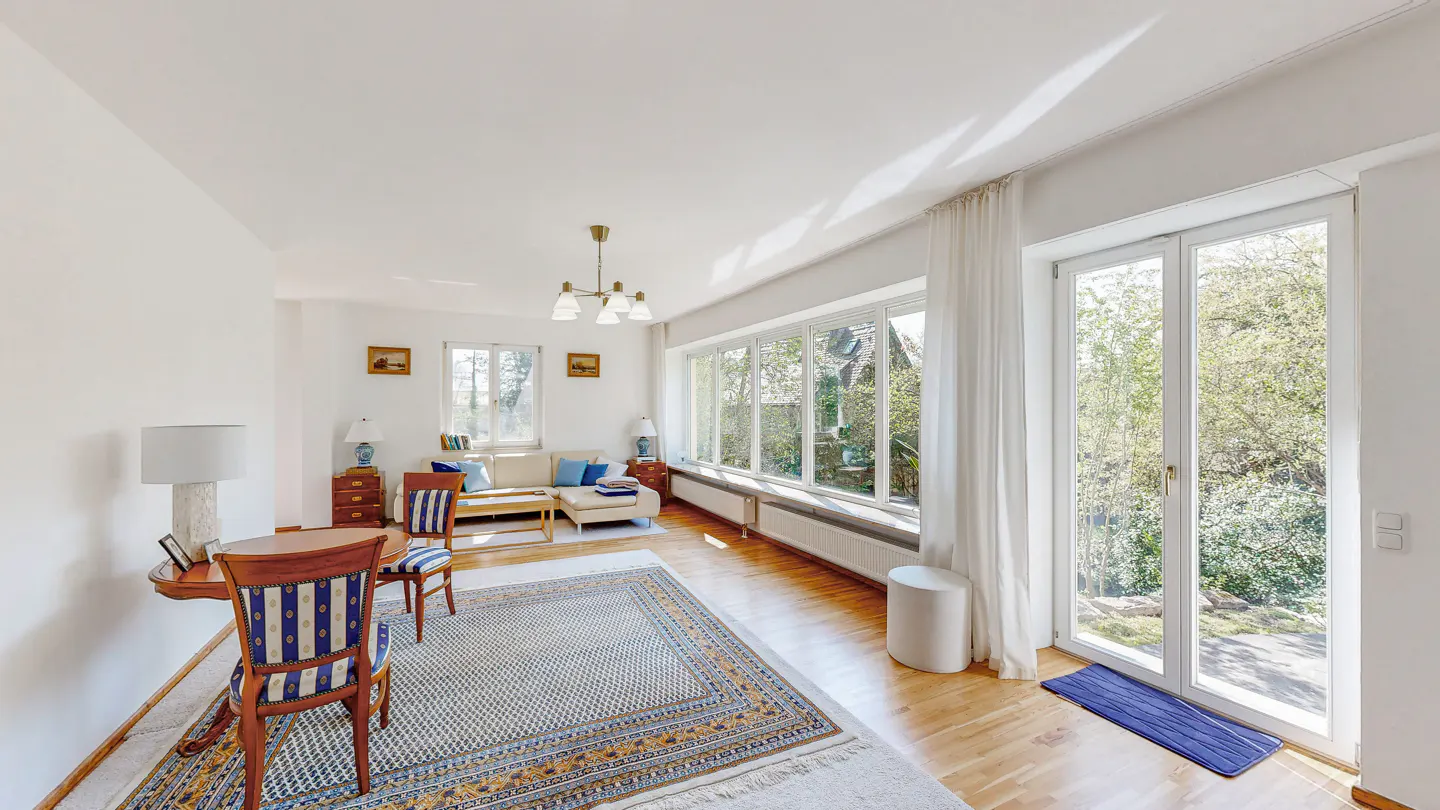 Bright living room with white walls, wood floors, and large windows. A blue and white rug sits under a table and chairs. A cream sofa is in the background.