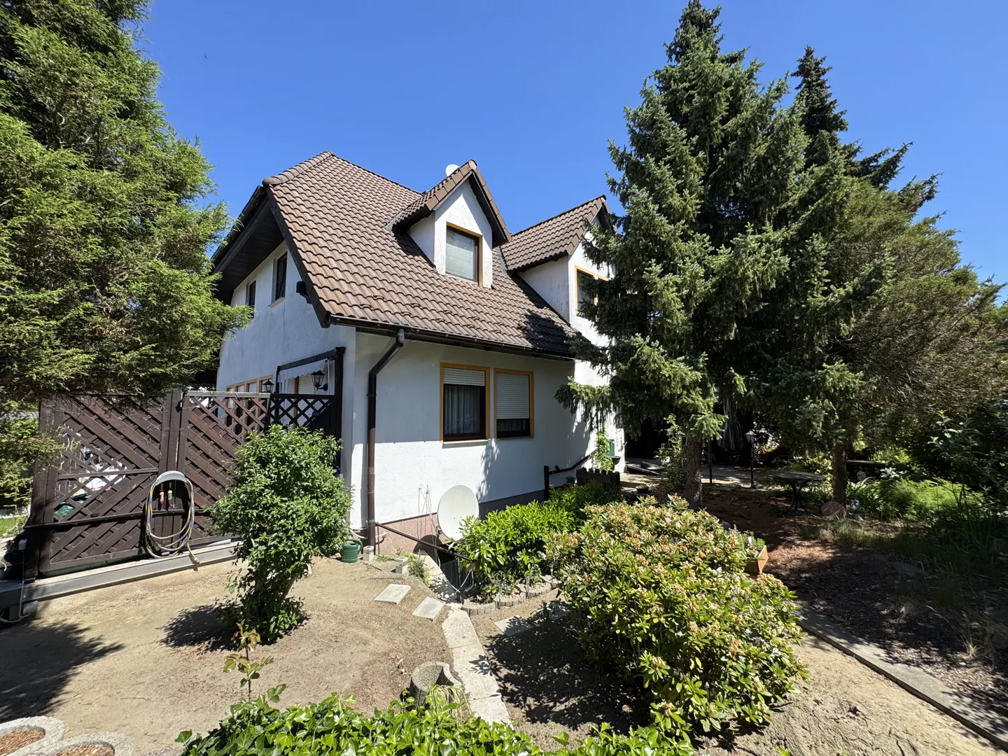 White house with a brown tiled roof, dormer windows, and a garden with large pine trees under a clear blue sky.