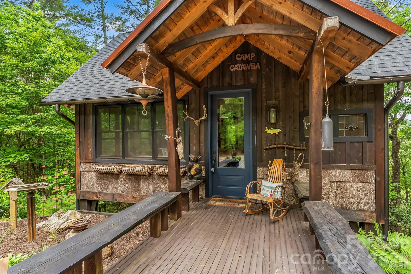 Rustic wooden cabin porch with a blue door, rocking chair, and "Camp Catawba" sign, surrounded by lush green forest.