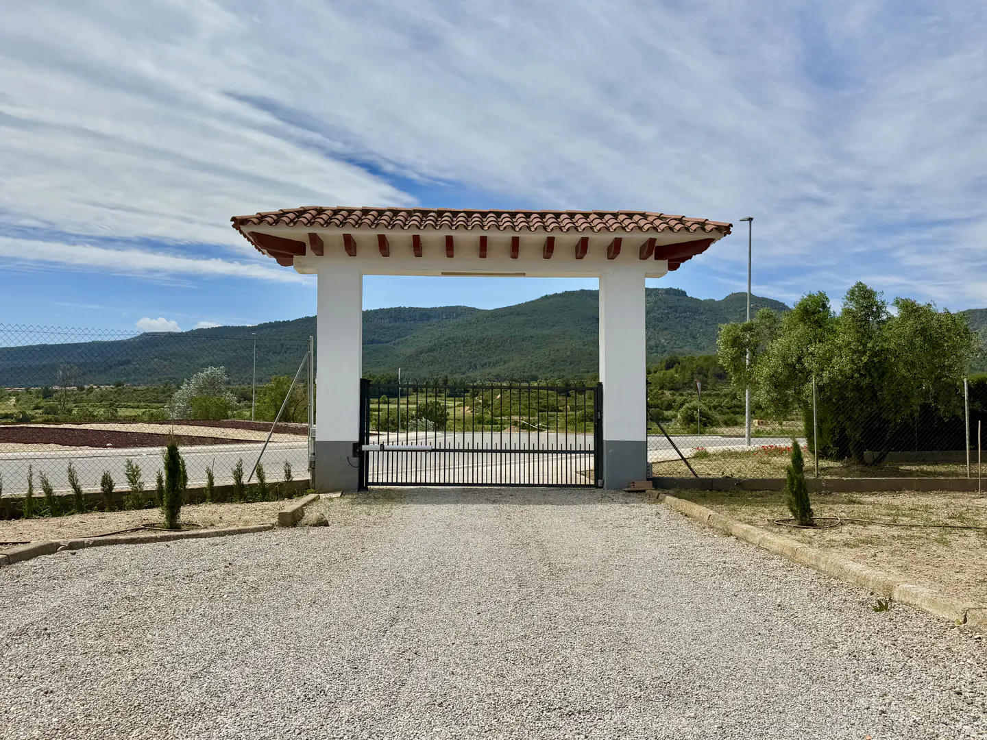 Gravel driveway with a black metal gate and white pillars topped with red tiles. Green hills are visible in the background.