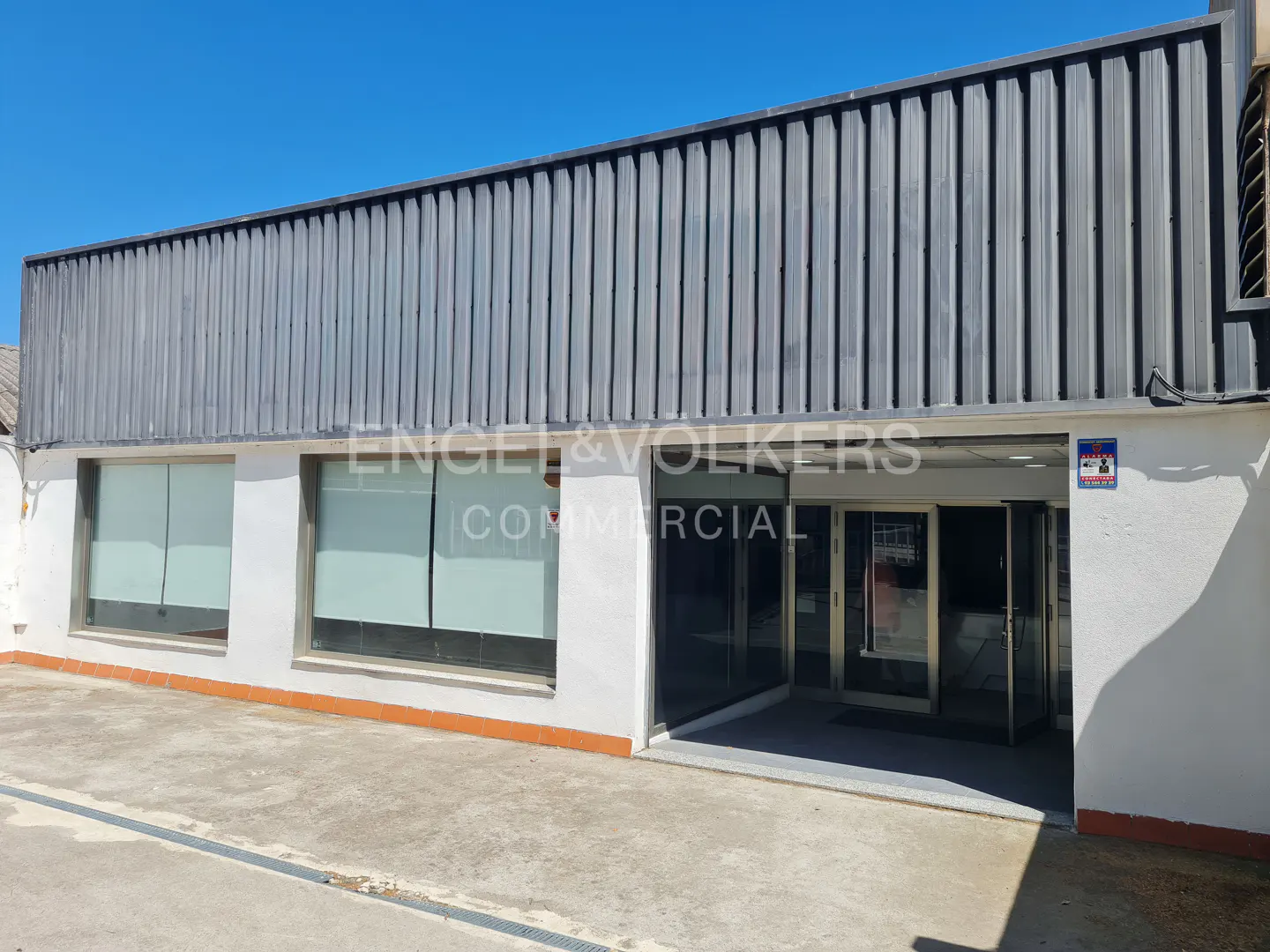 Exterior of a commercial building with white walls, large windows, and a dark grey corrugated metal roof under a blue sky.