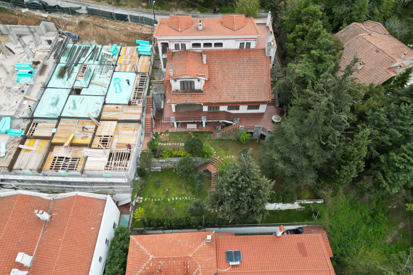 Aerial view of a white house with a red tile roof, a green garden, and a construction site next door.