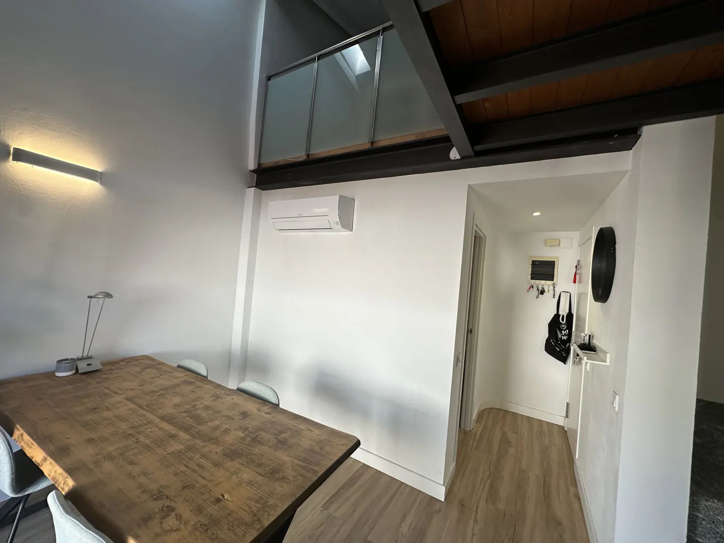 Interior view of a modern apartment with a wooden table, chairs, and a hallway leading to a white-walled area with a chalkboard and bag.
