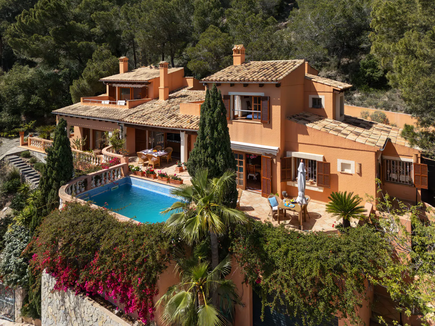 Aerial view of a terracotta-colored villa with a tiled roof, surrounded by lush greenery and a blue swimming pool. Outdoor dining and seating areas are visible.