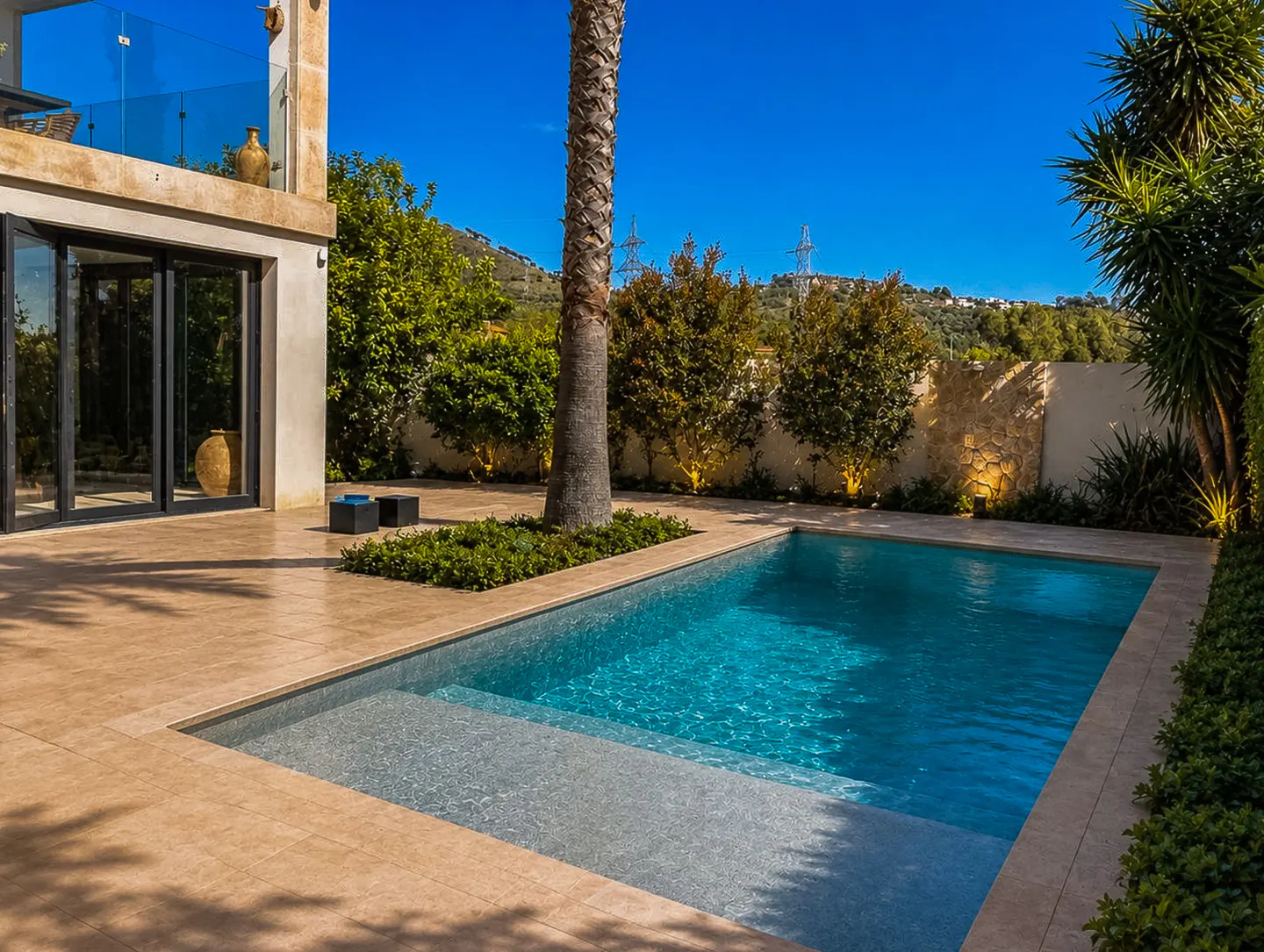 A rectangular pool with turquoise water is surrounded by a tan tiled patio and lush greenery. A modern building stands nearby.