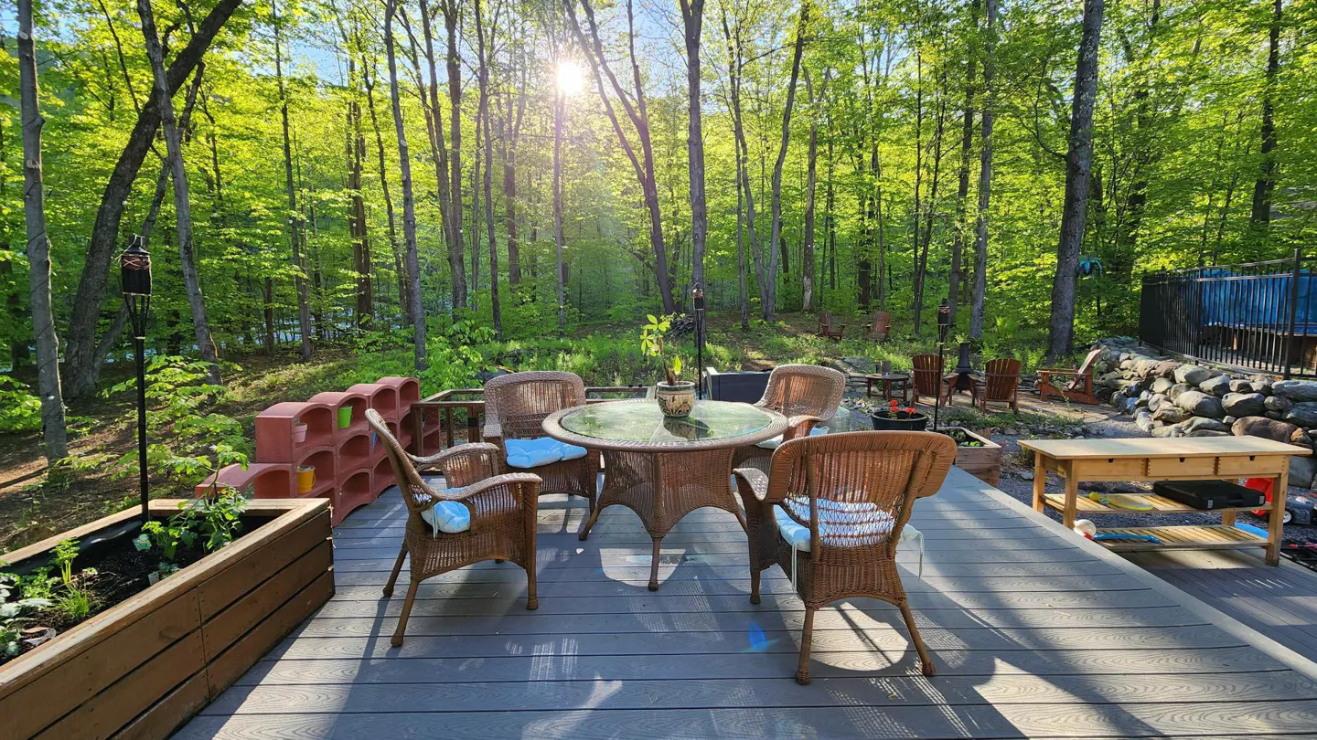 Outdoor patio with wicker table and chairs on a gray deck, surrounded by lush green trees and sunlight.