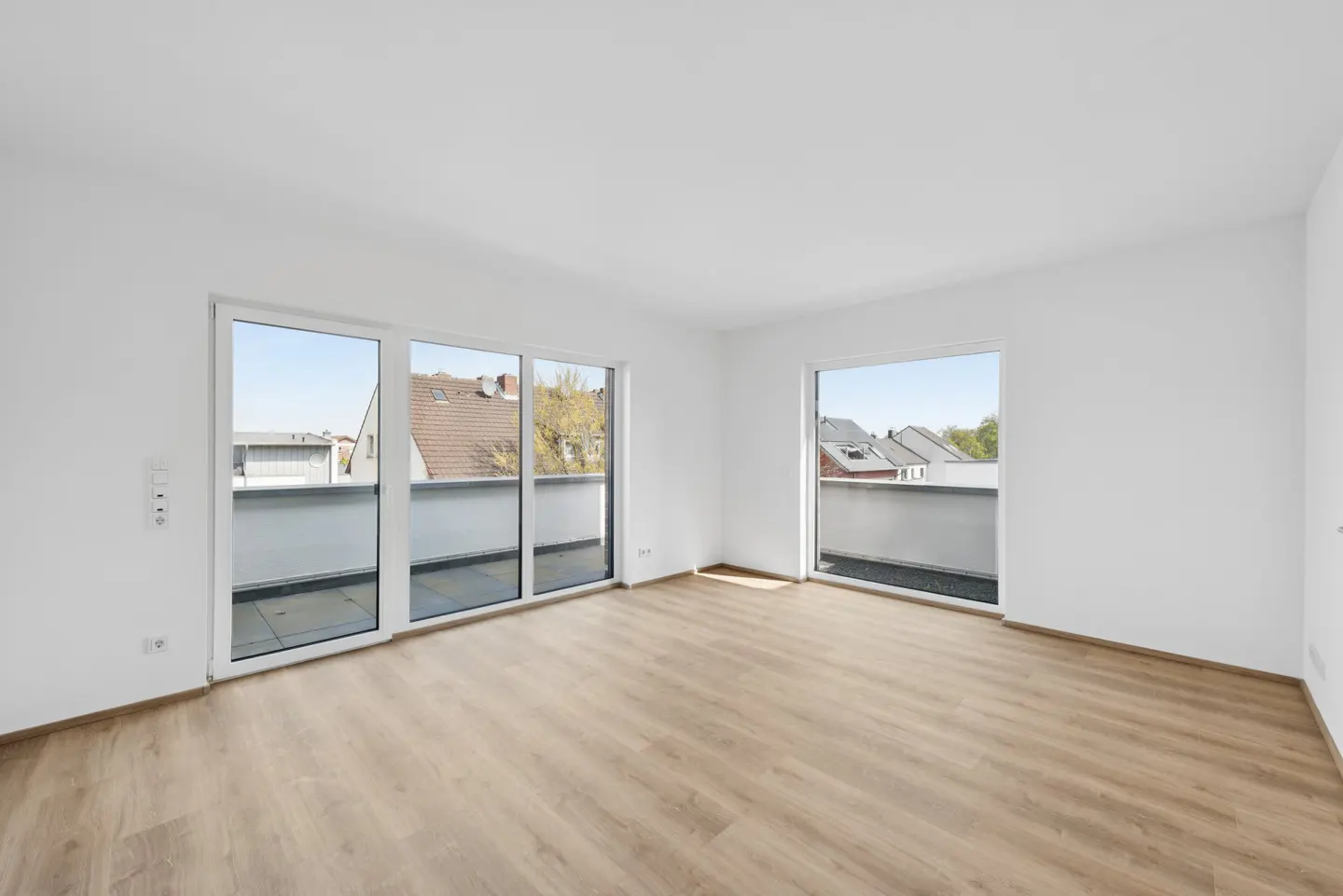 Bright, empty room with light wood floors and white walls. Large sliding glass doors lead to balconies with neighborhood views.
