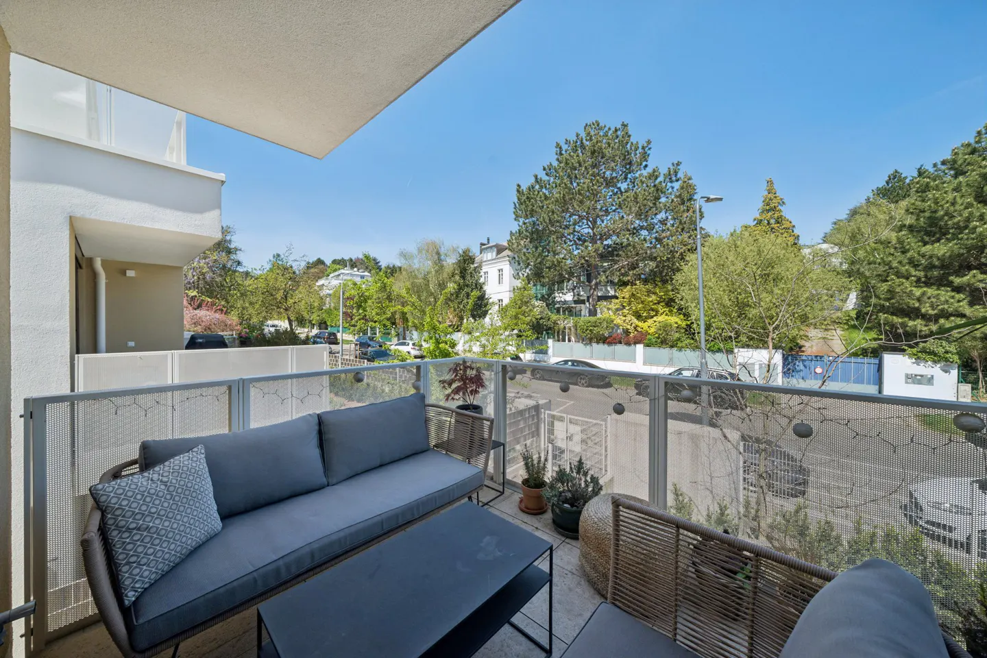 Balcony with gray sofa, black table, and woven chairs. Glass railing overlooks a street with trees and houses under a blue sky.