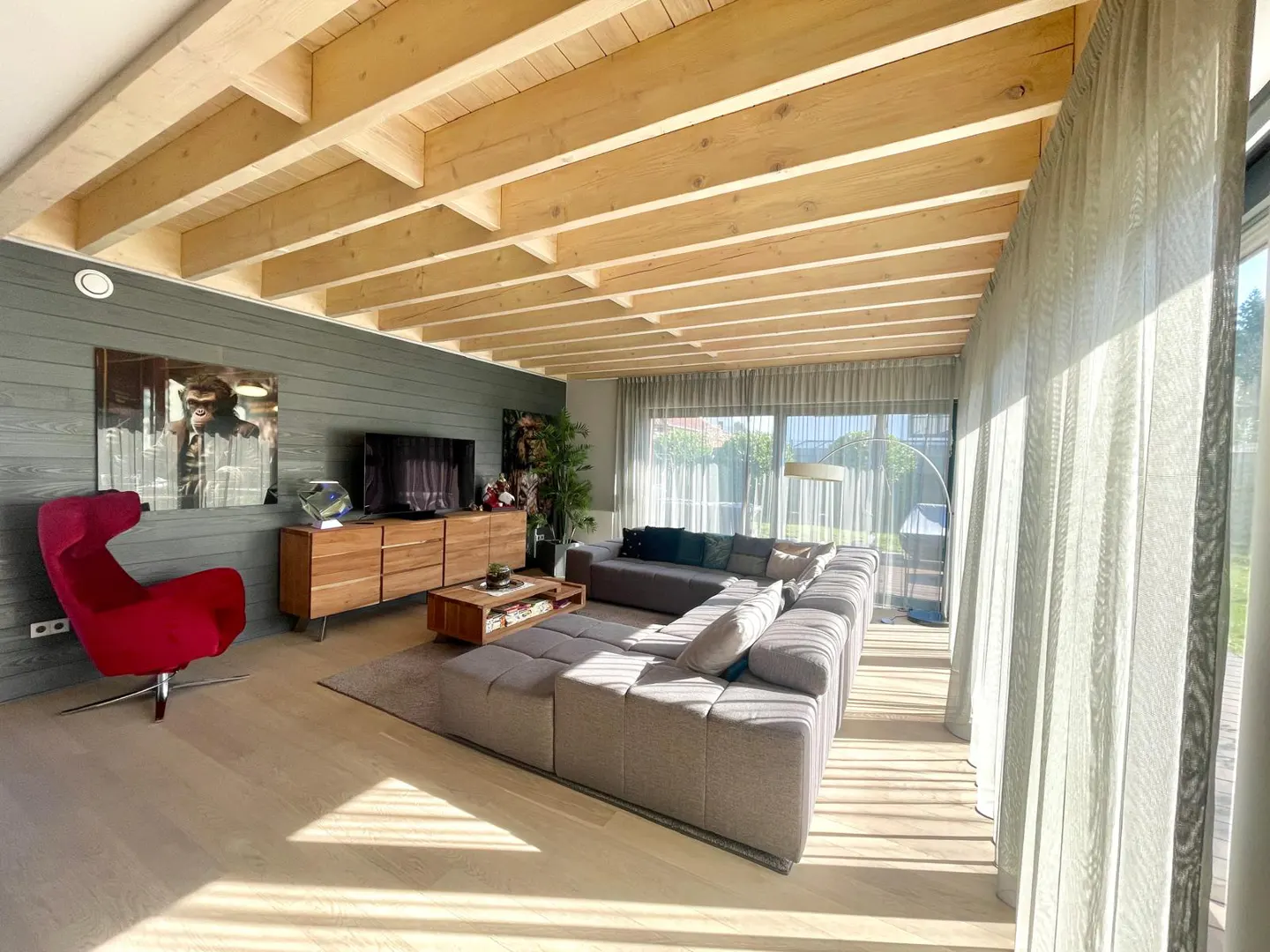 Bright living room with wood-beamed ceiling, gray walls, and light wood floors. A large gray sectional sofa faces a wooden media console and TV. A red chair adds a pop of color.