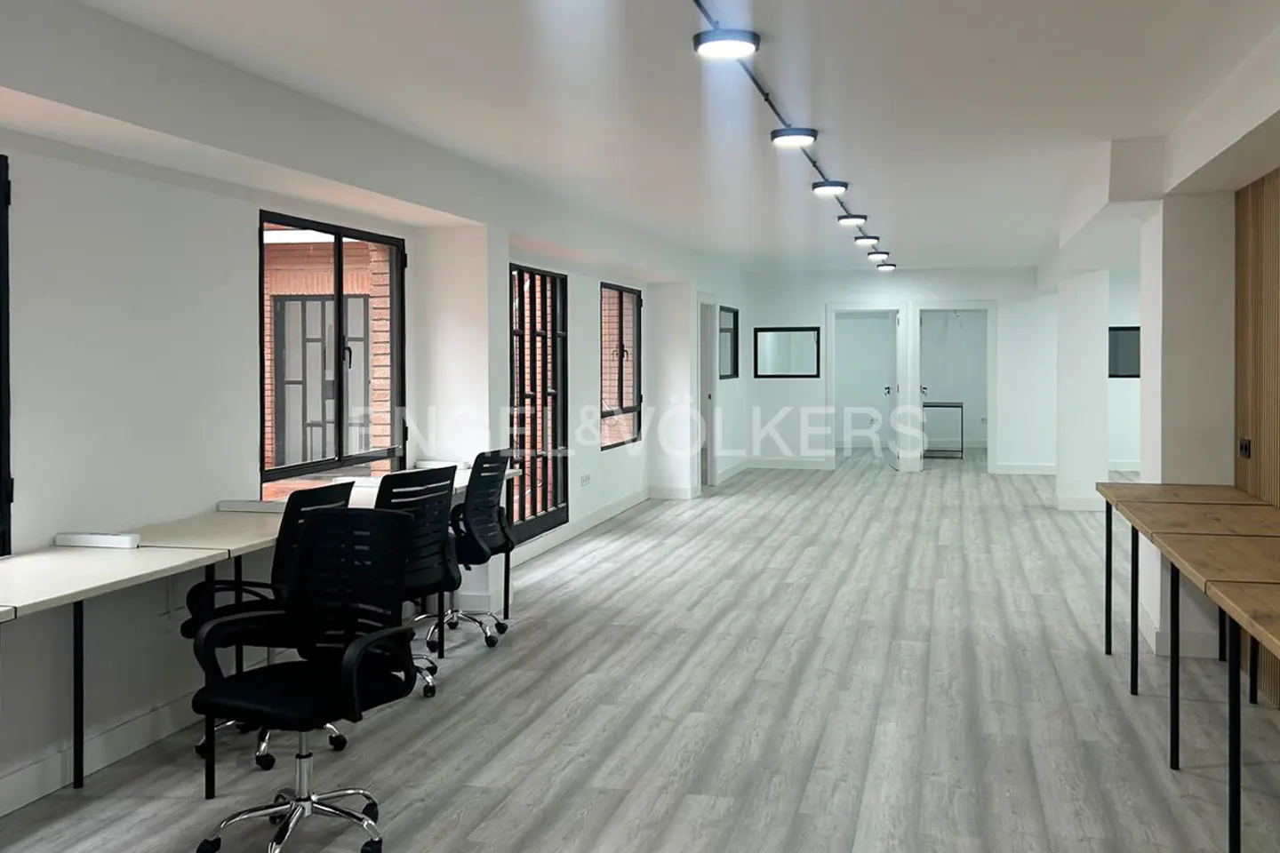 Bright, empty office space with white walls, gray wood floors, and black-framed windows. Desks with black chairs line one wall.